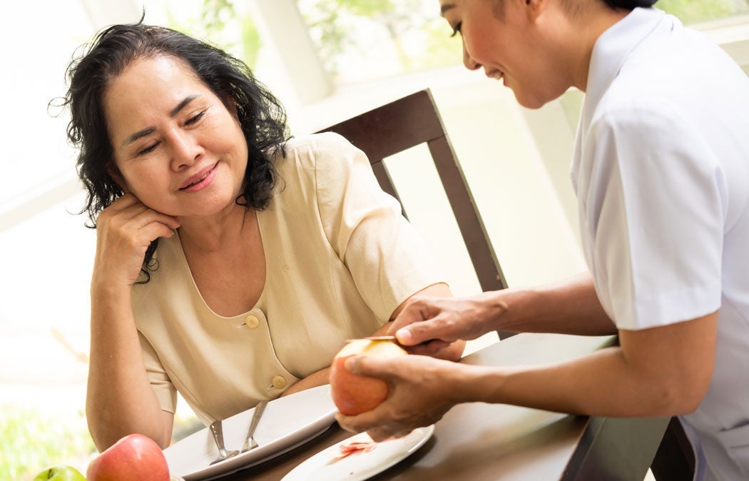 Caregiver assisting an older person with preparing food at a table.