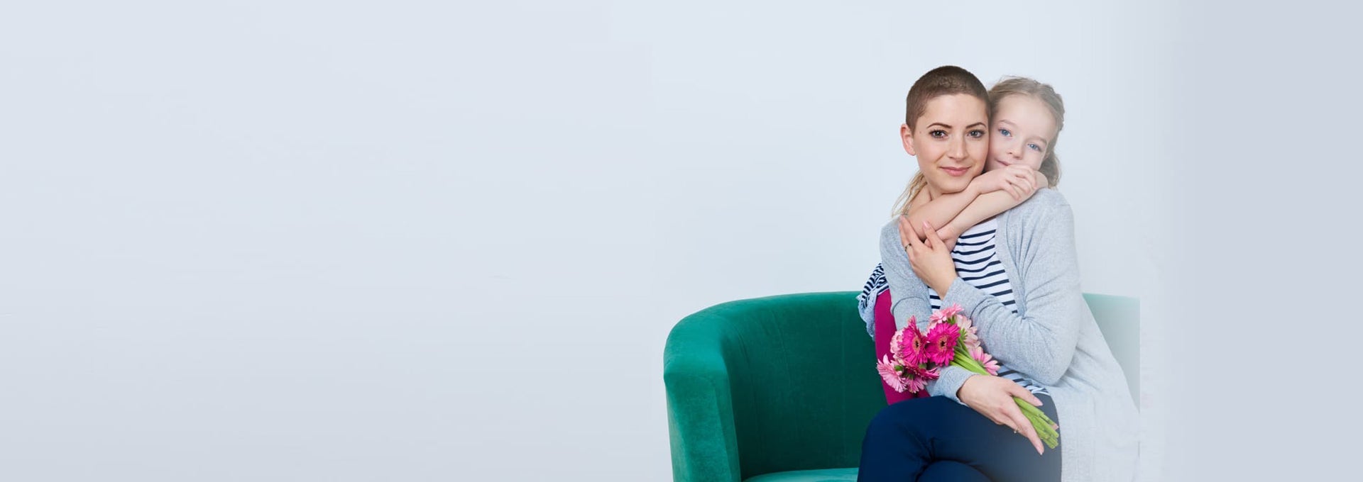 Person sitting on a green chair holding flowers while a child hugs from behind against a light background.