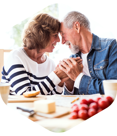 Two older adults holding hands and leaning close together at a kitchen table with food items.